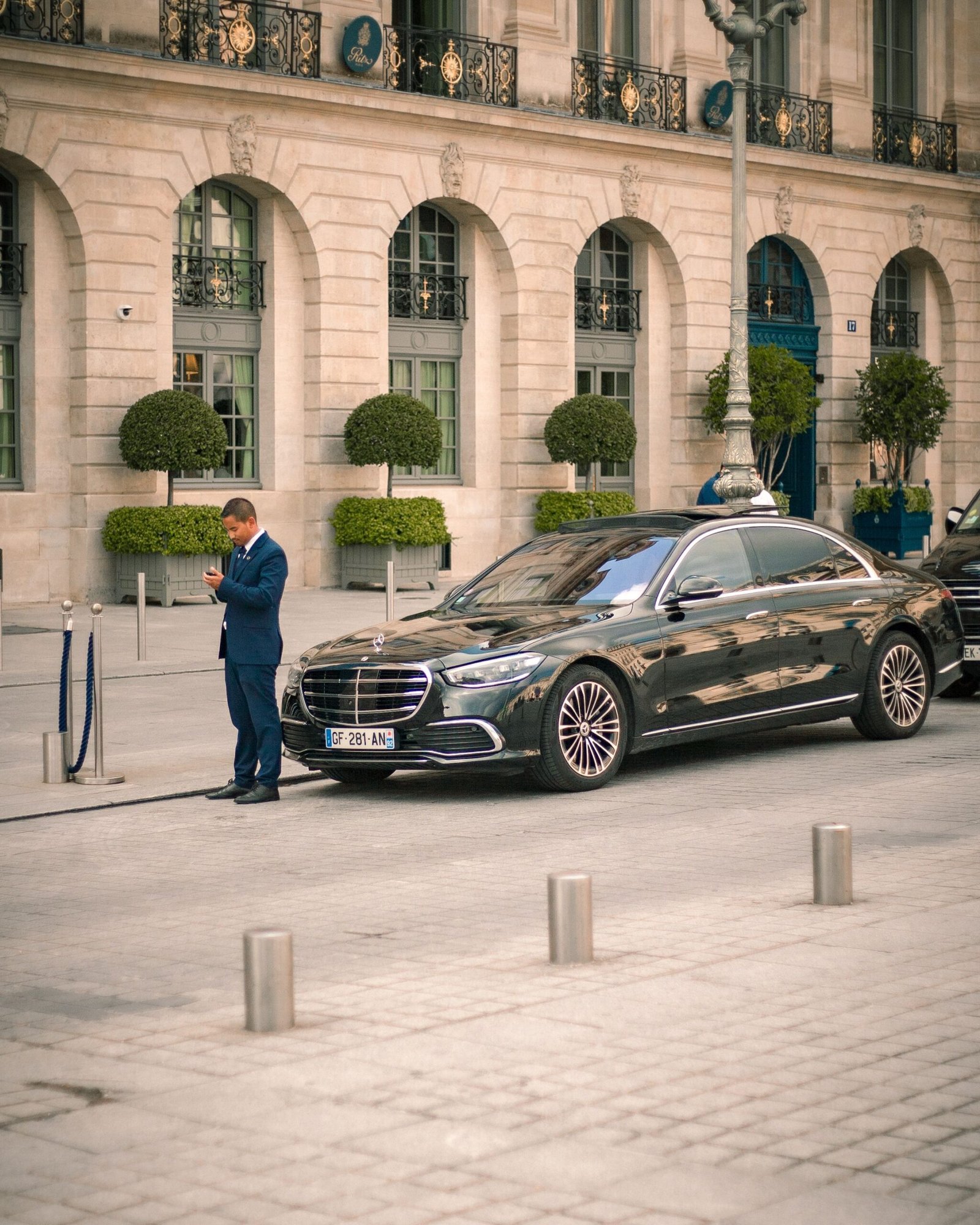 Businessman using a smartphone next to a luxury car parked on a European city street.
